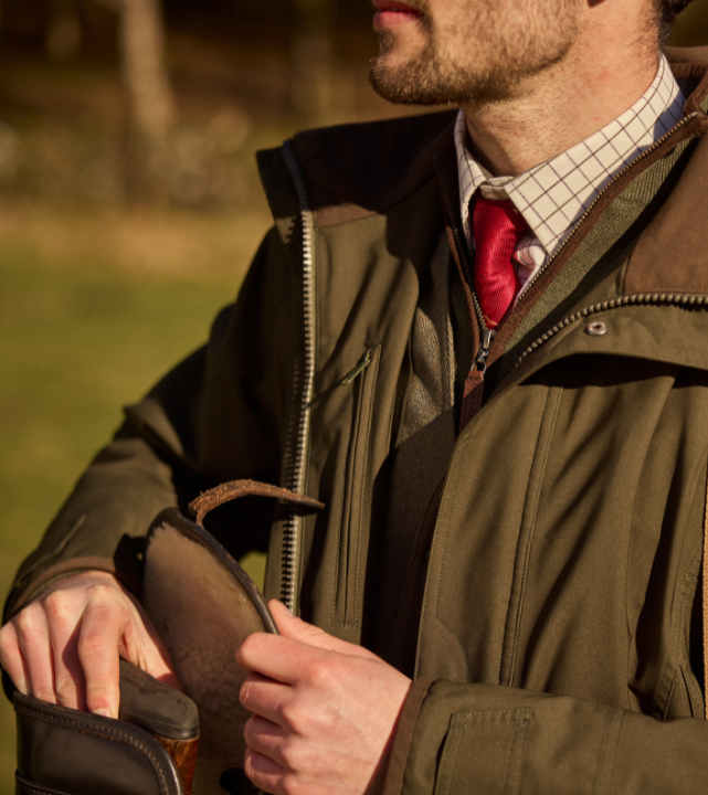 Person wearing a woodland green outdoor jacket, opening a front pocket while standing outdoors.