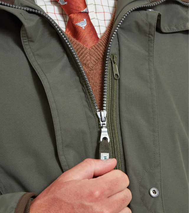 Close-up of the open front of a forest green ultralight coat with a dual-zip fastening and silver zip pulls, with a hand gripping the lower zip pull. A rust knit jumper and a check shirt with an orange ptarmigan-print tie are visible underneath.