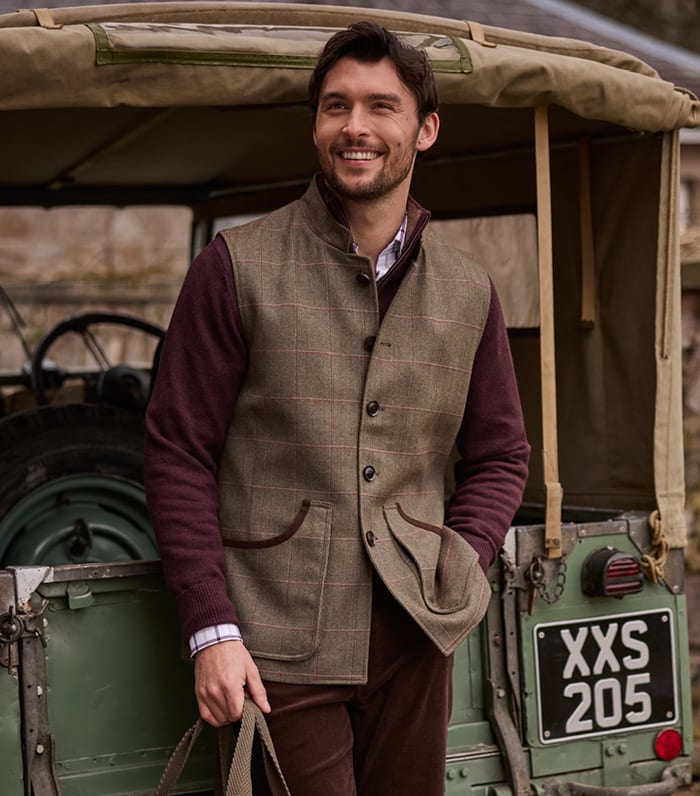 A man wearing a brown tweed waistcoat and brown trousers, standing in front of a green vintage vehicle.