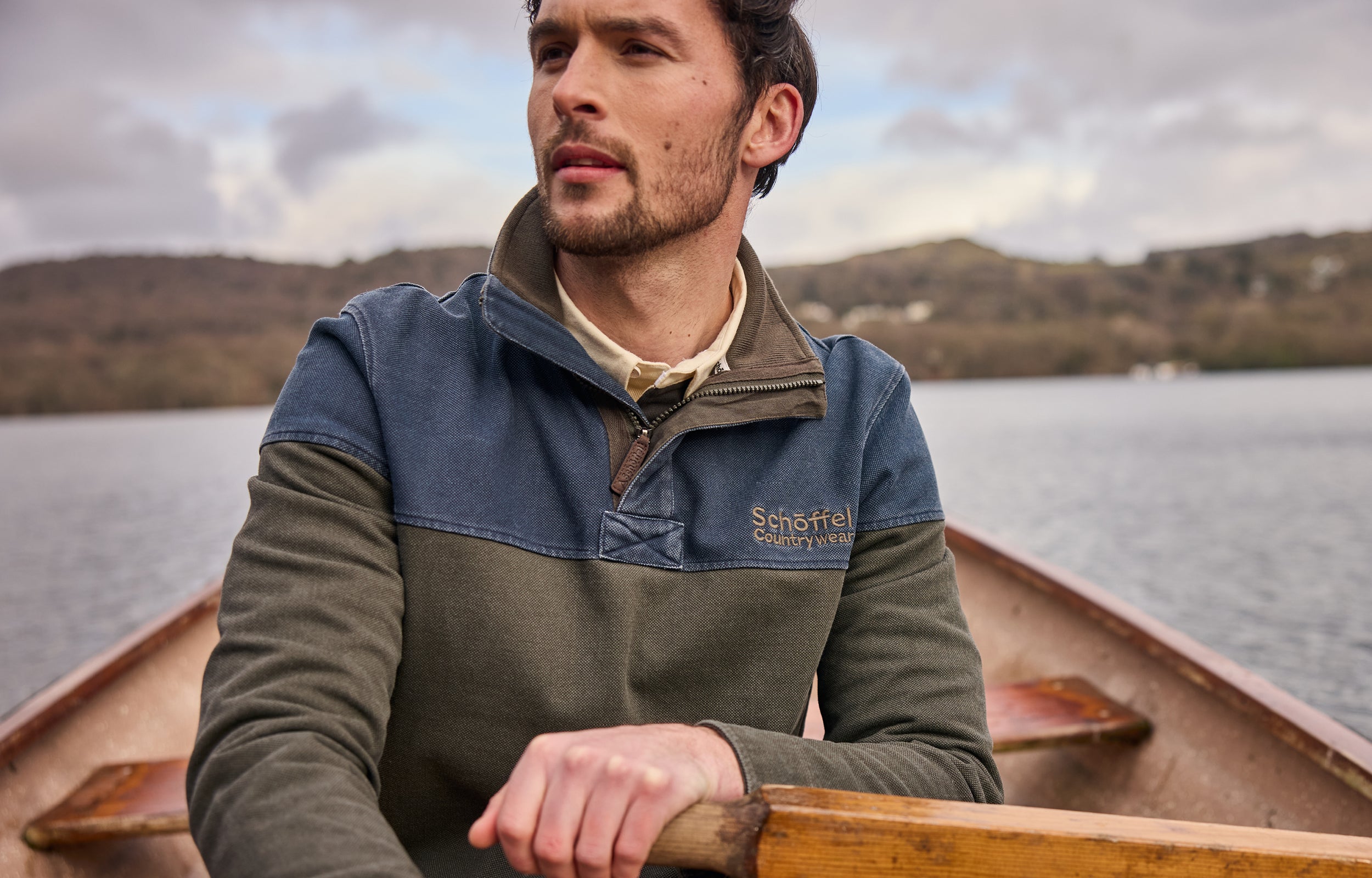 Man in a branded sweatshirt rowing a boat on a lake with mountains in the background