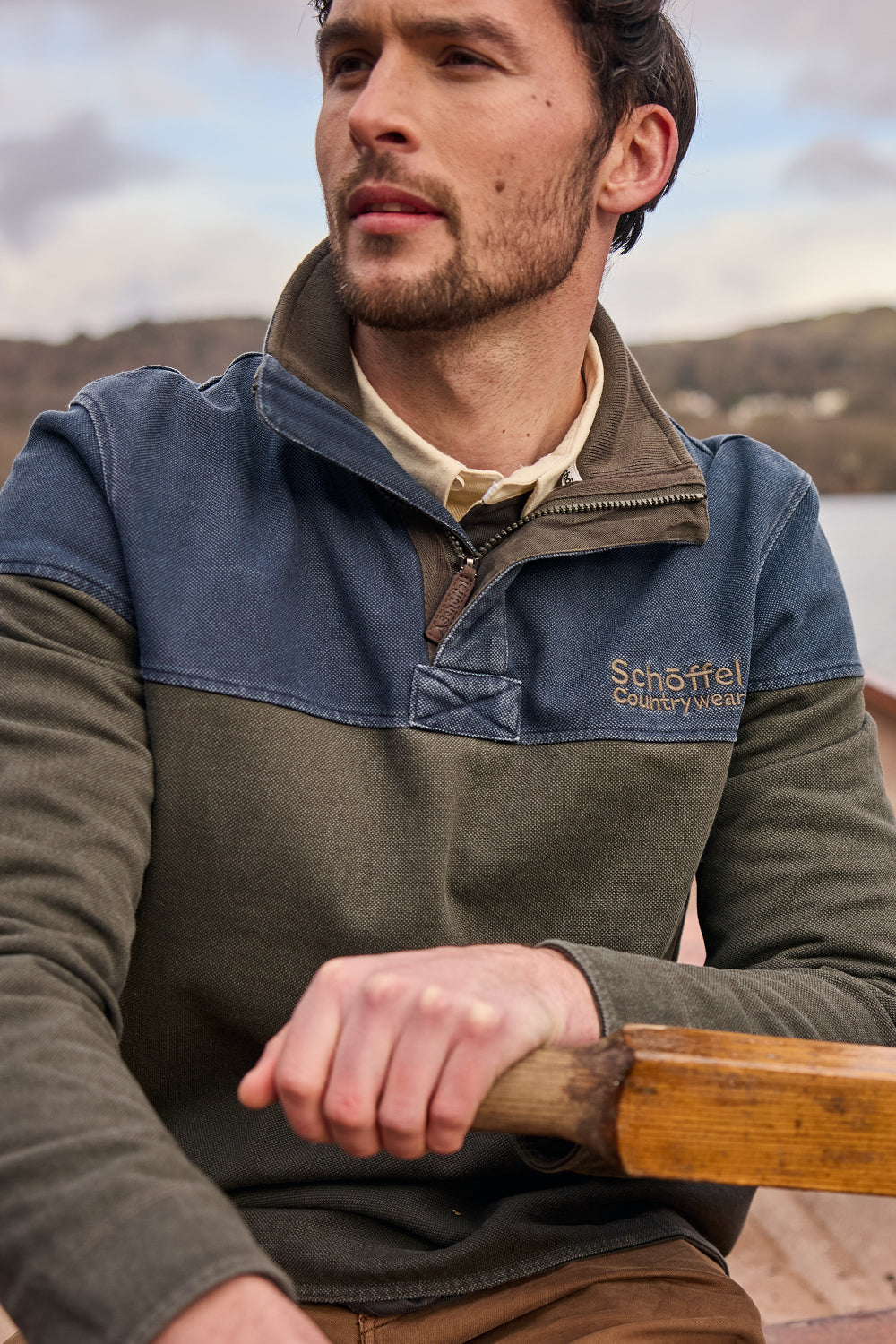 Man in a branded sweatshirt rowing a boat on a lake with mountains in the background