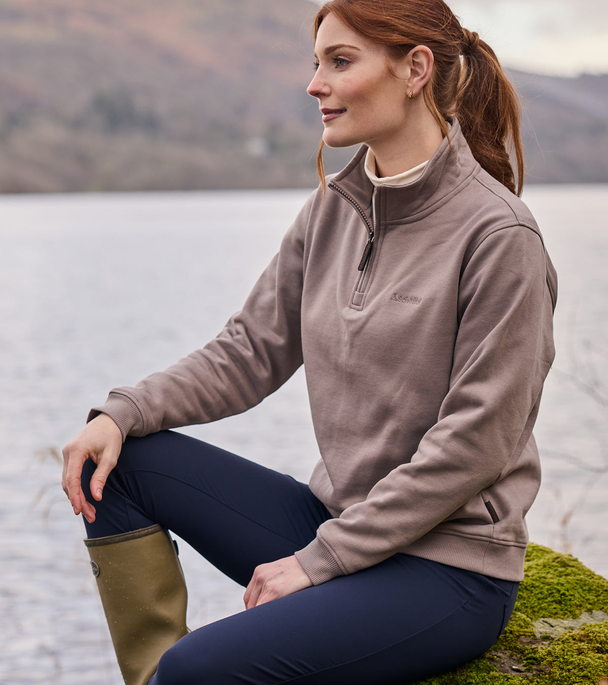 Woman wearing a brown sweatshirt sitting by a lake with mountains in the background