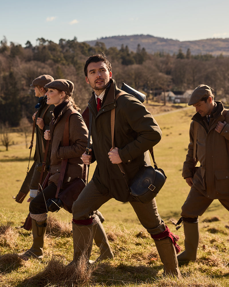Four people in outdoor clothing walking through a field with trees and mountains in the background.