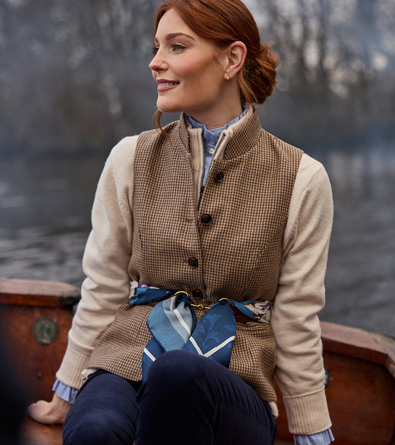 Woman sitting on a boat wearing a tweed vest and beige sweatshirt with a blurred water background
