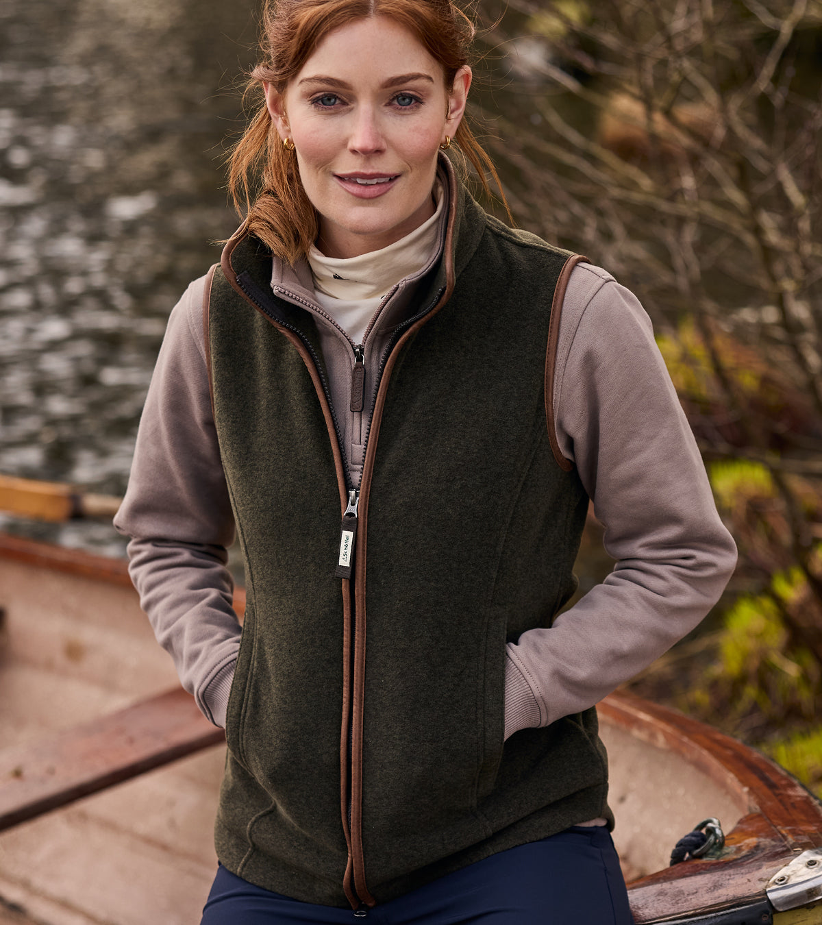 Woman wearing a green gilet over a brown sweatshirt by a lake.