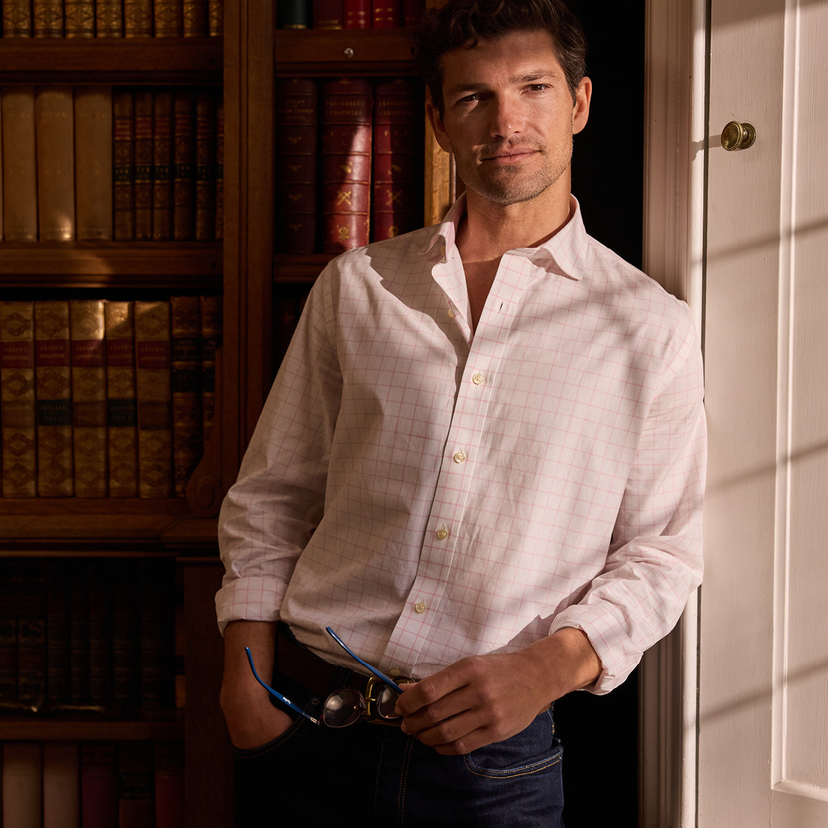 Man in a white and pink checked shirt standing in a room with bookshelves