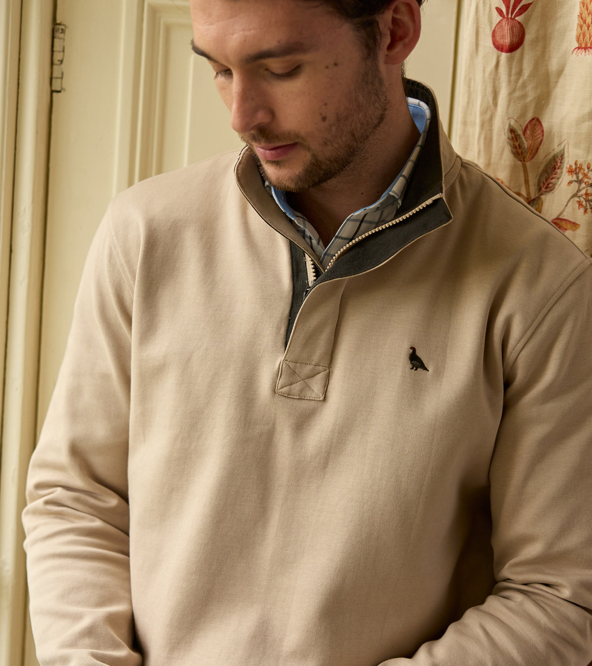 Man wearing a beige sweatshirt with a small logo, standing indoors.