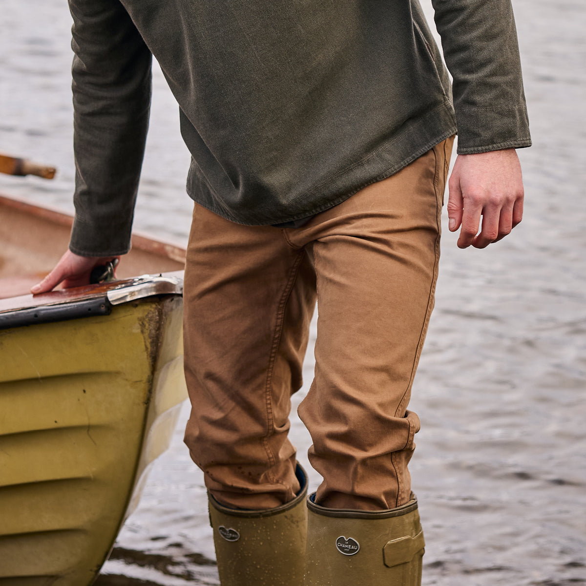 Person wearing brown trousers and green sweatshirt standing next to a yellow canoe by a body of water.