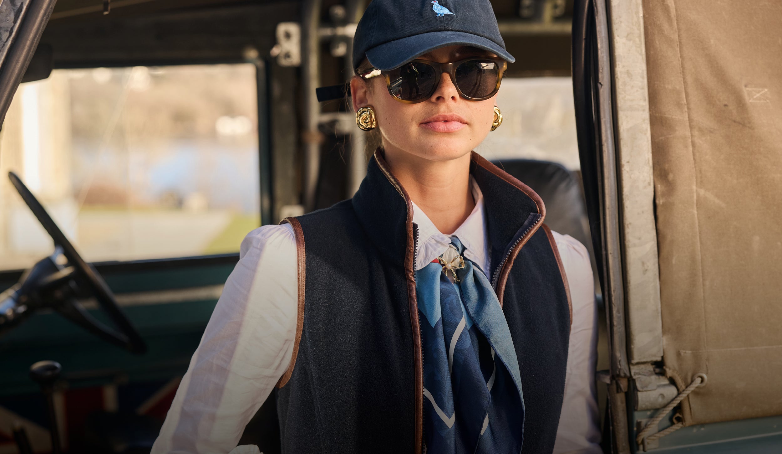 Person wearing a navy gilet, white shirt, blue scarf, sunglasses, and a navy cap.