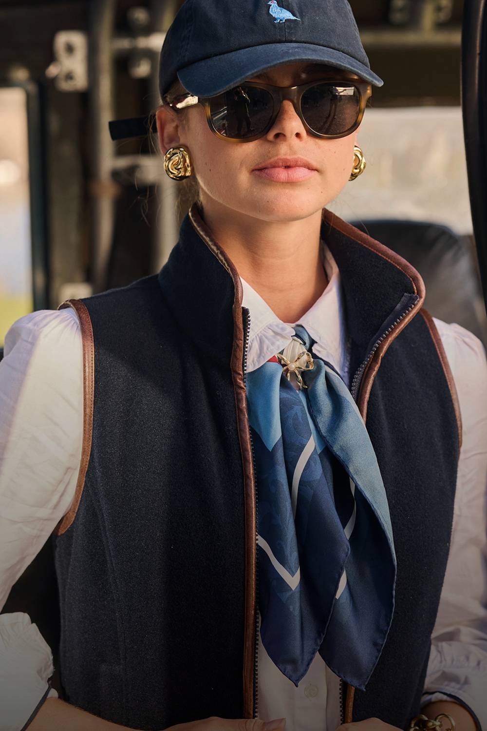 Person wearing a navy gilet, white shirt, blue scarf, sunglasses, and a navy cap.