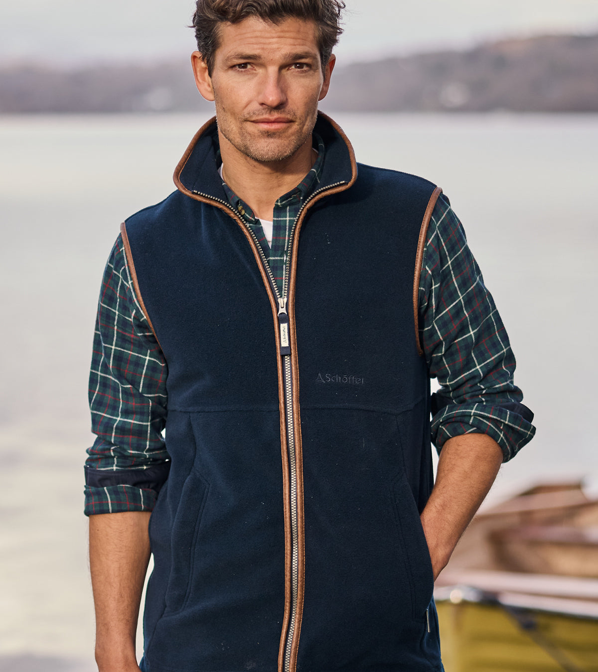 Man wearing a navy fleece gilet with a checked shirt, standing by a lake