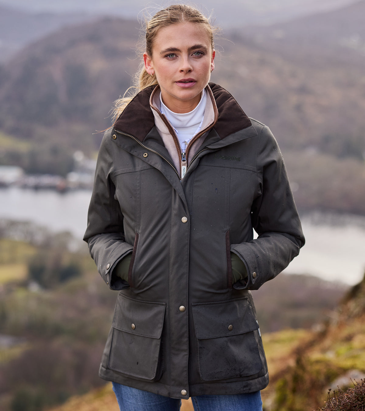 Woman wearing a green jacket in a scenic outdoor setting with mountains and water.