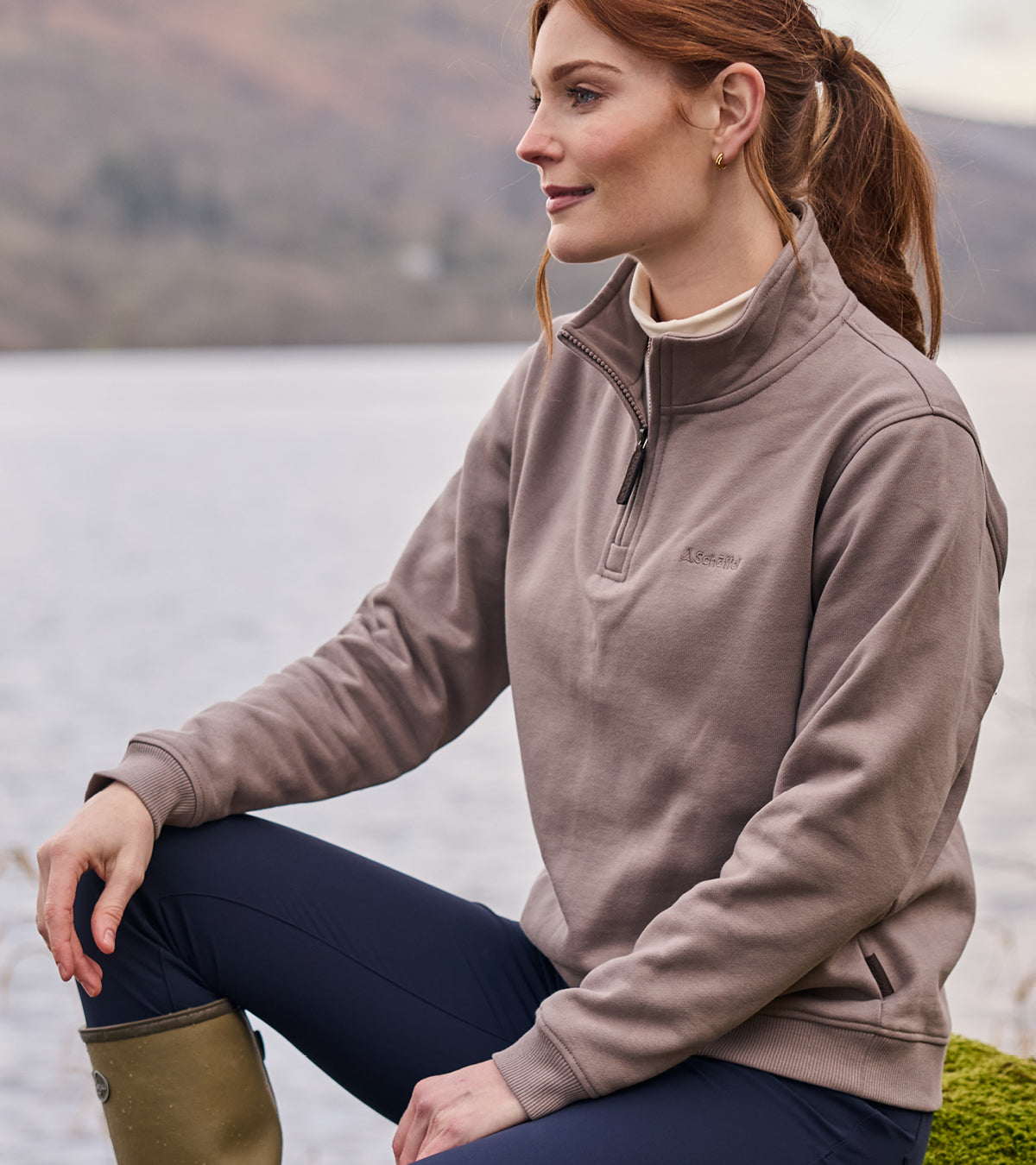 Woman wearing a brown quarter zip sweatshirt sitting by a lake