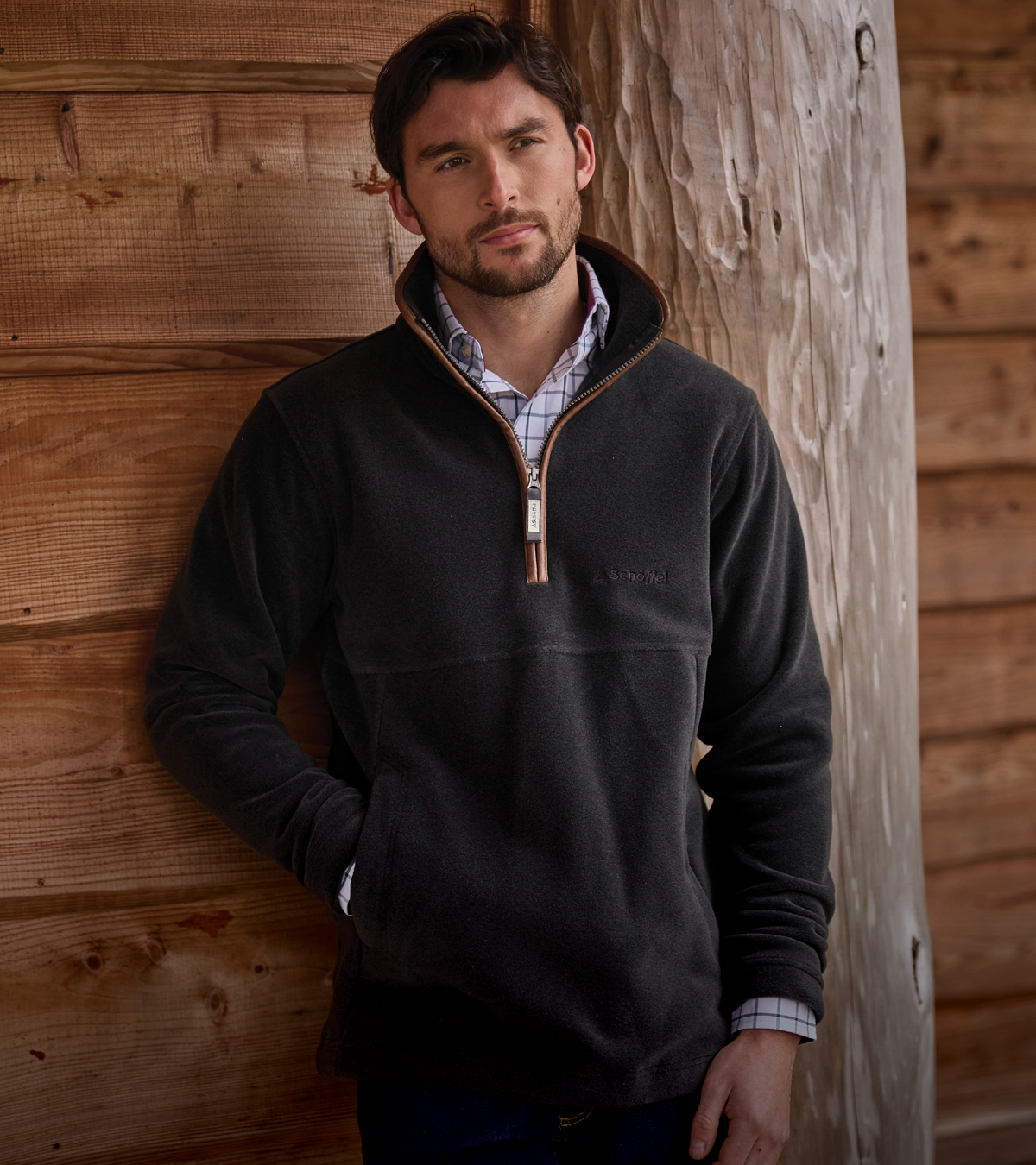 A man wearing a dark charcoal gray quarter-zip fleece pullover with a brown zipper trim, standing against a rustic wooden wall.