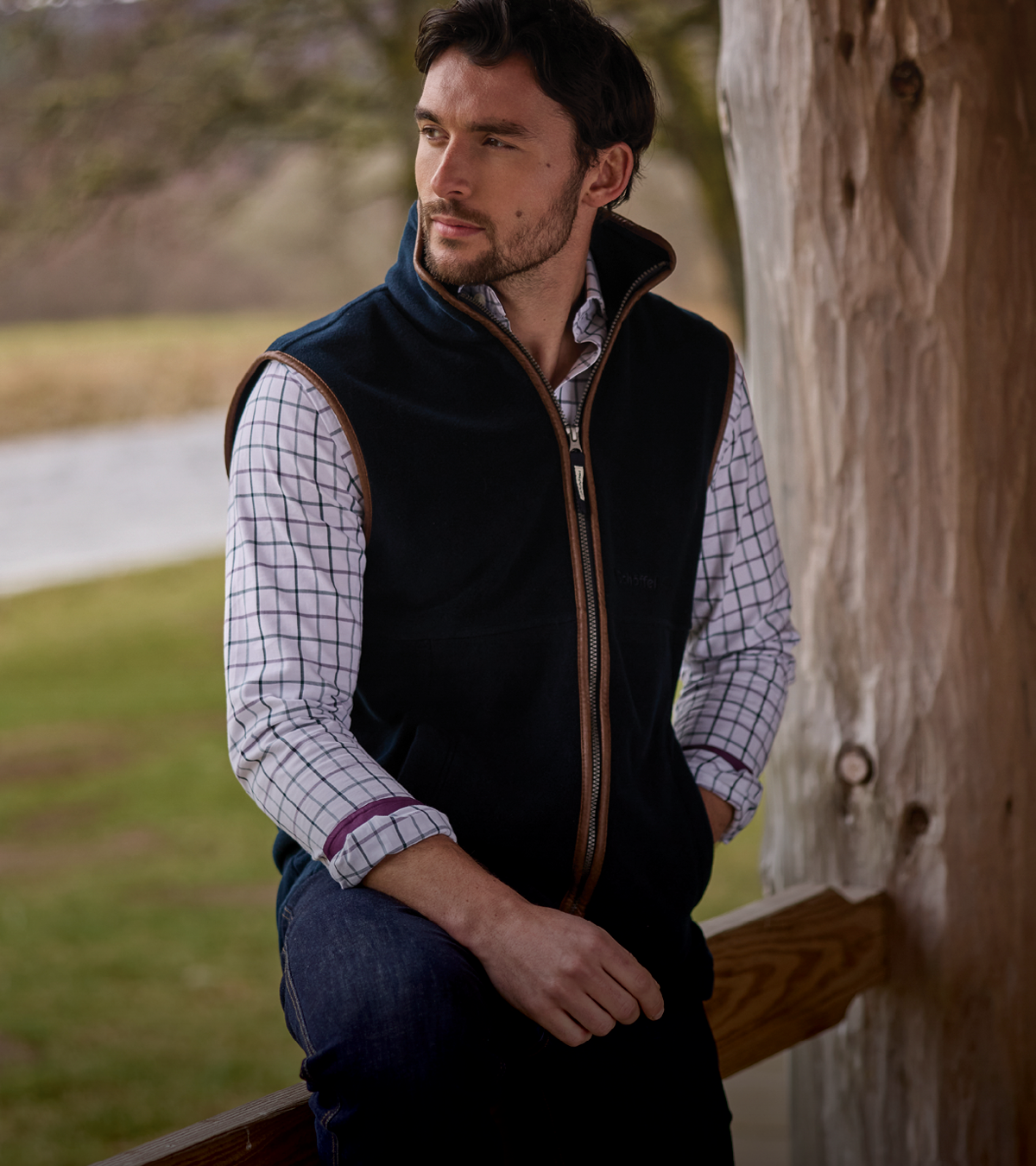 A man wearing a navy fleece zip-front gilet over a white and navy checked shirt, sitting outdoors next to a tree trunk.