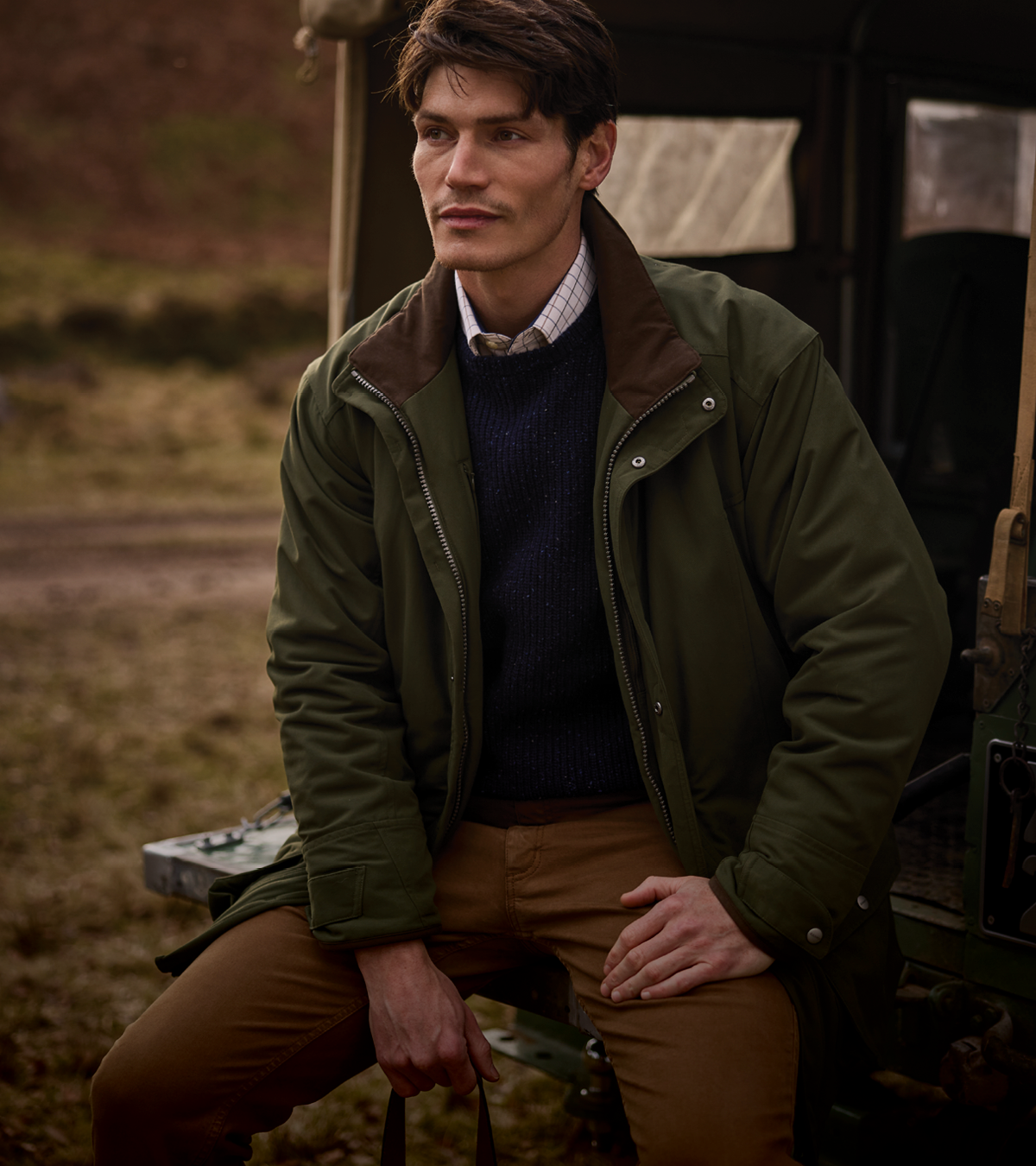 A man wearing an olive green jacket with brown collar panels, a navy jumper, and brown trousers, seated outdoors next to a vehicle.