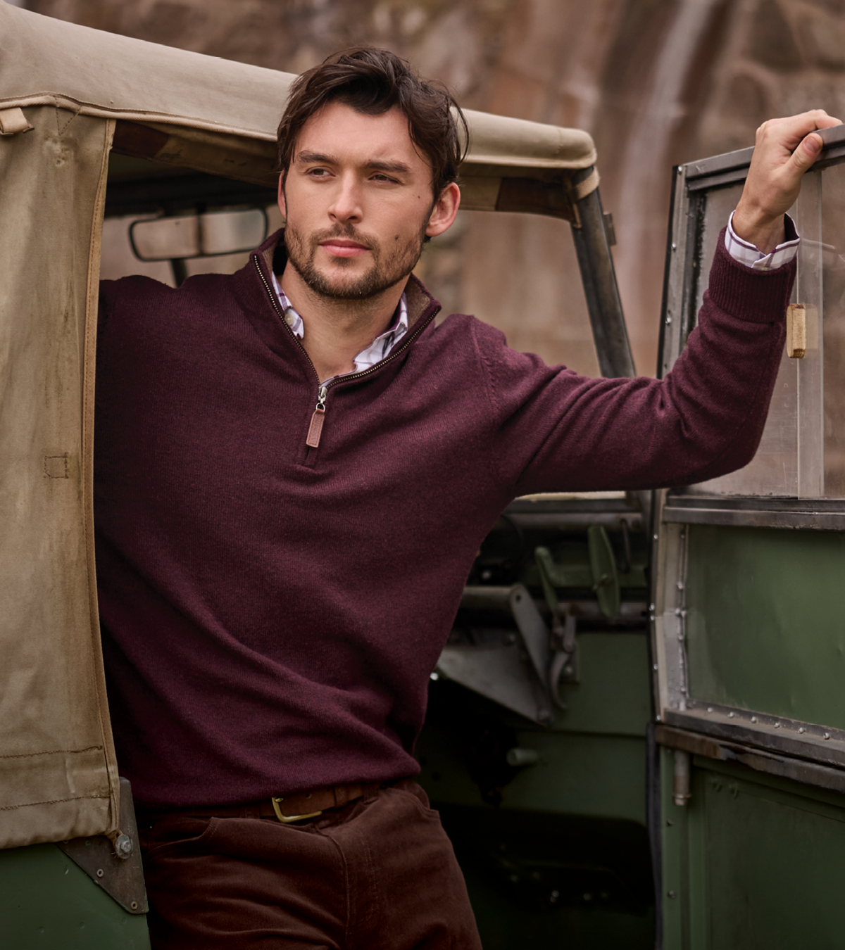 A man wearing a burgundy quarter-zip jumper and brown trousers leans out of an olive green vintage vehicle, with a stone wall in the background.