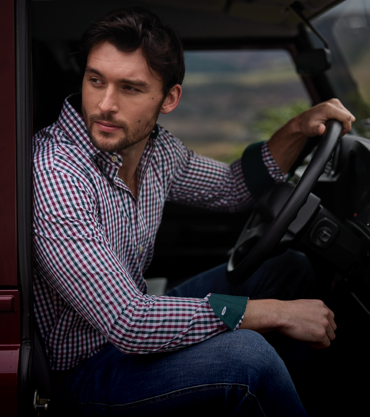 A man wearing a red, white, and green gingham long-sleeve button-down shirt and blue jeans sits in the driver's seat of a vehicle, looking out the window.