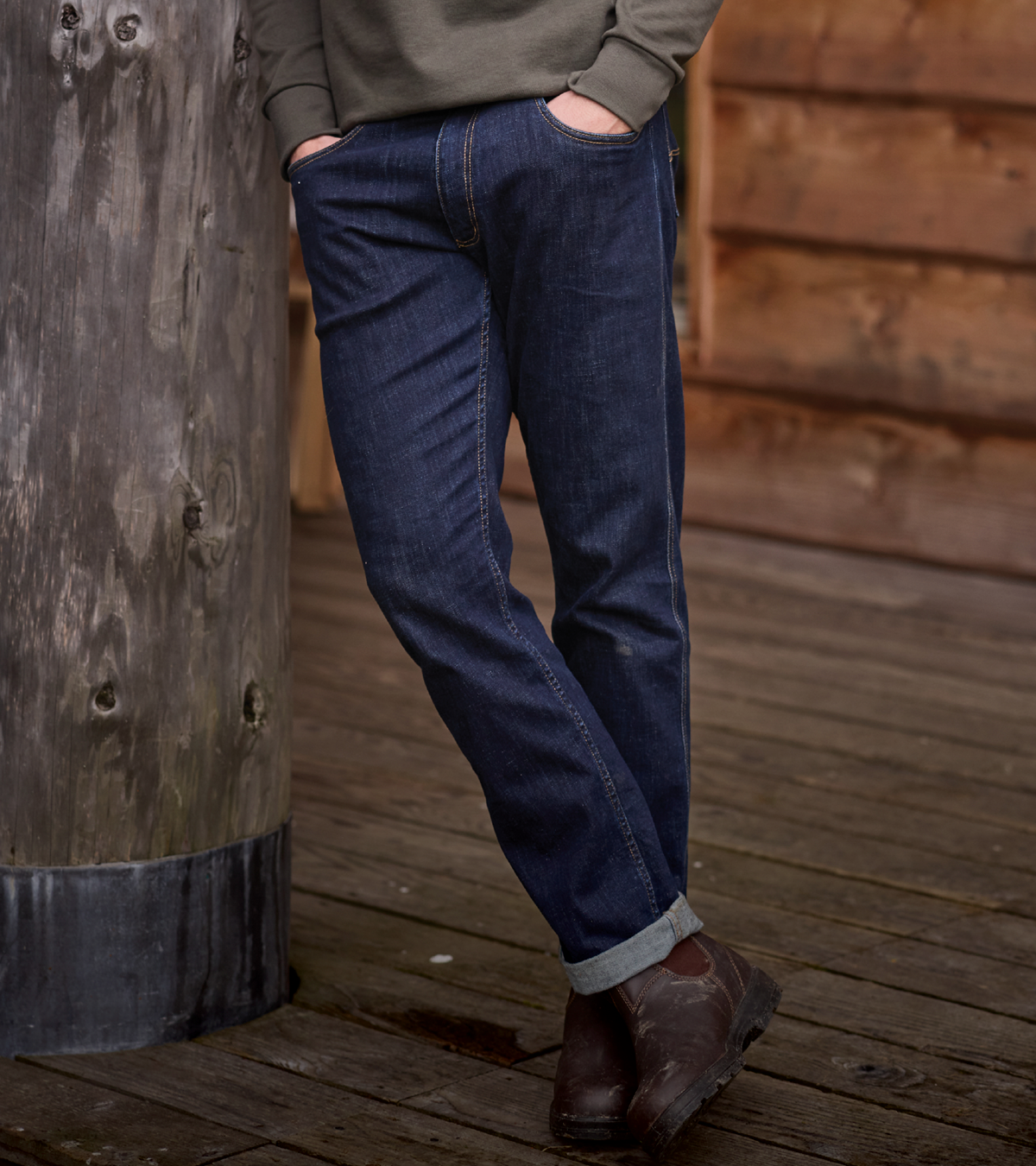 A person wearing dark blue straight-leg jeans with a cuffed hem, paired with a grey sweatshirt and brown leather boots, standing on a wooden deck next to a rustic wooden post.