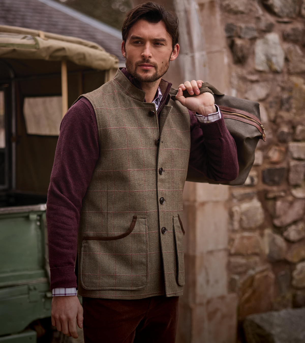 A man wearing a brown tweed waistcoat over a burgundy jumper, standing outdoors next to a stone wall and carrying a brown leather bag.