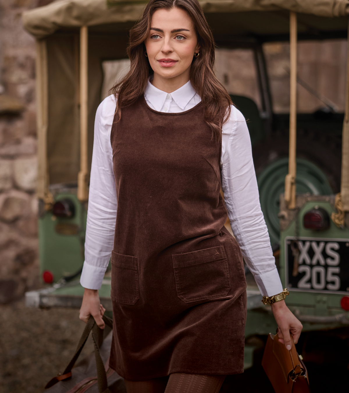 A woman wearing a brown corduroy pinafore dress over a white collared shirt, standing in front of a vintage green vehicle.