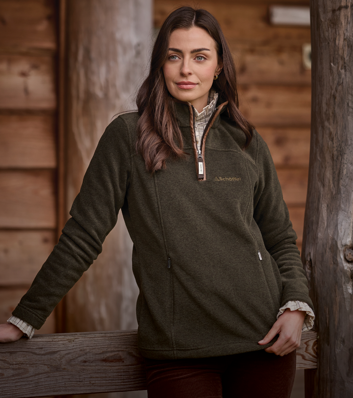 A woman wearing an olive green quarter-zip fleece pullover, styled over a patterned shirt and dark brown trousers, leaning against a rustic wooden fence in front of a wooden cabin wall.