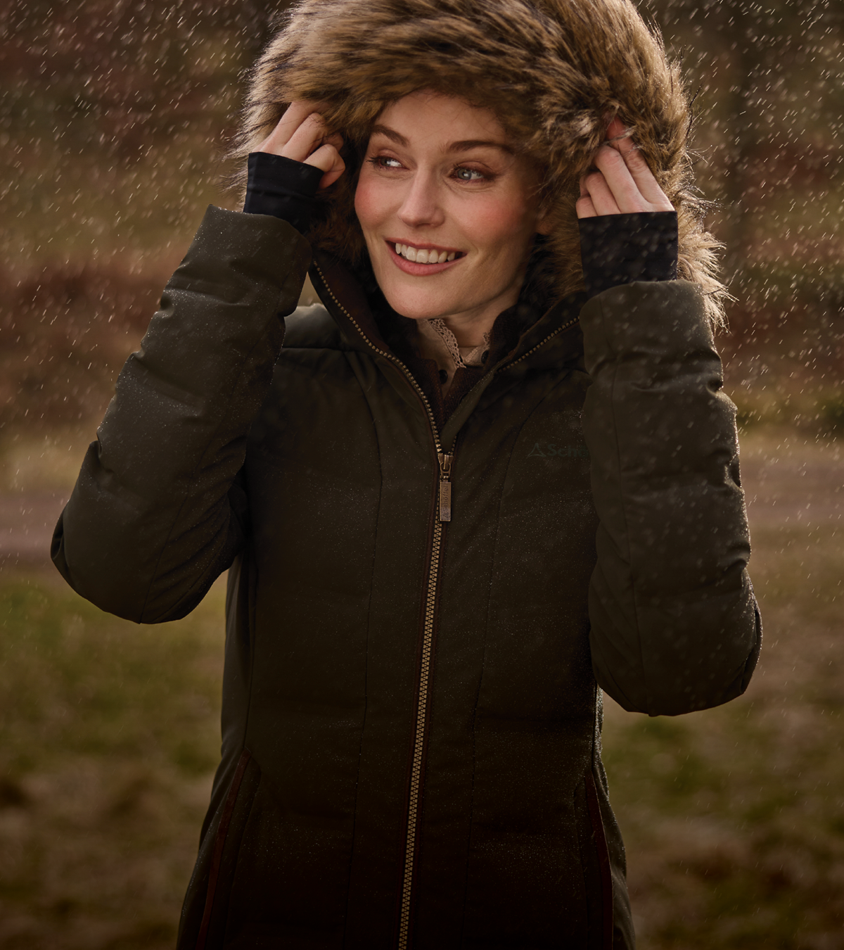A woman smiling in a dark olive green puffer jacket with a large brown faux-fur hood, holding the hood up in a rainy outdoor setting.