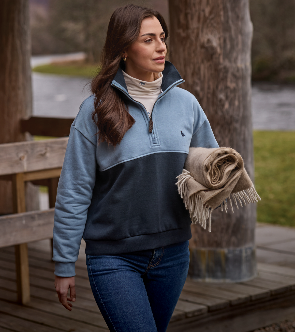 A woman walking outdoors on a wooden walkway wearing a color-blocked quarter-zip sweatshirt in light blue and navy, paired with a cream turtleneck and blue jeans, while holding a rolled beige fringed blanket.