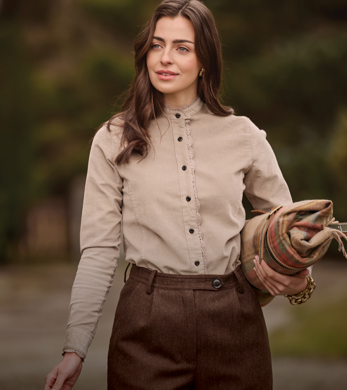 A woman wearing a beige long-sleeve button-front blouse with lace trim and brown trousers, holding a rolled multicolor knit blanket outdoors against a blurred natural background.