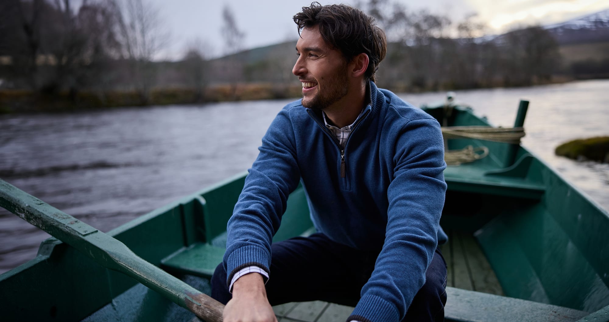 A man wearing a blue quarter-zip jumper sits in a green boat on a river, looking off-camera with a smile.