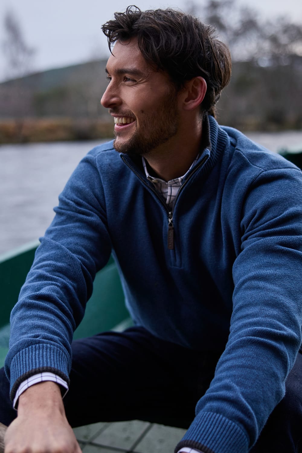 A man wearing a blue quarter-zip jumper sits in a boat on a lake, looking to the side with a smile.