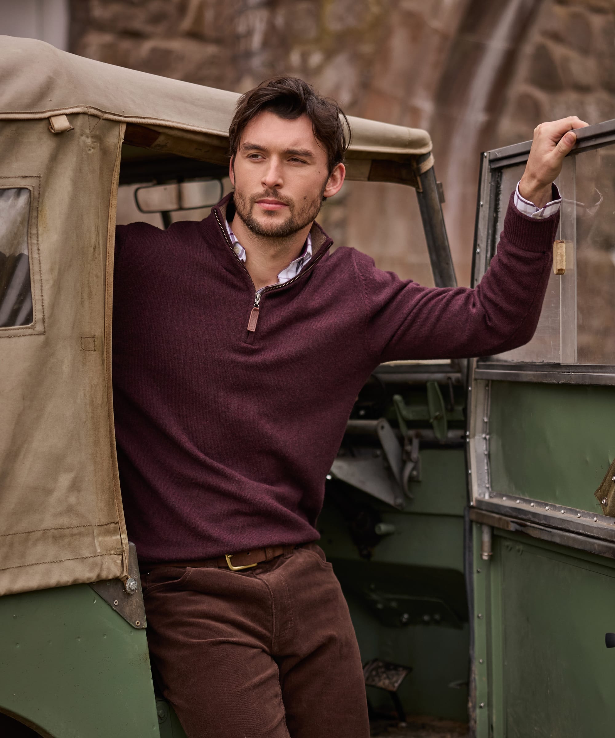 A man wearing a burgundy quarter-zip jumper and brown cord trousers leans against an olive green vintage vehicle outdoors.