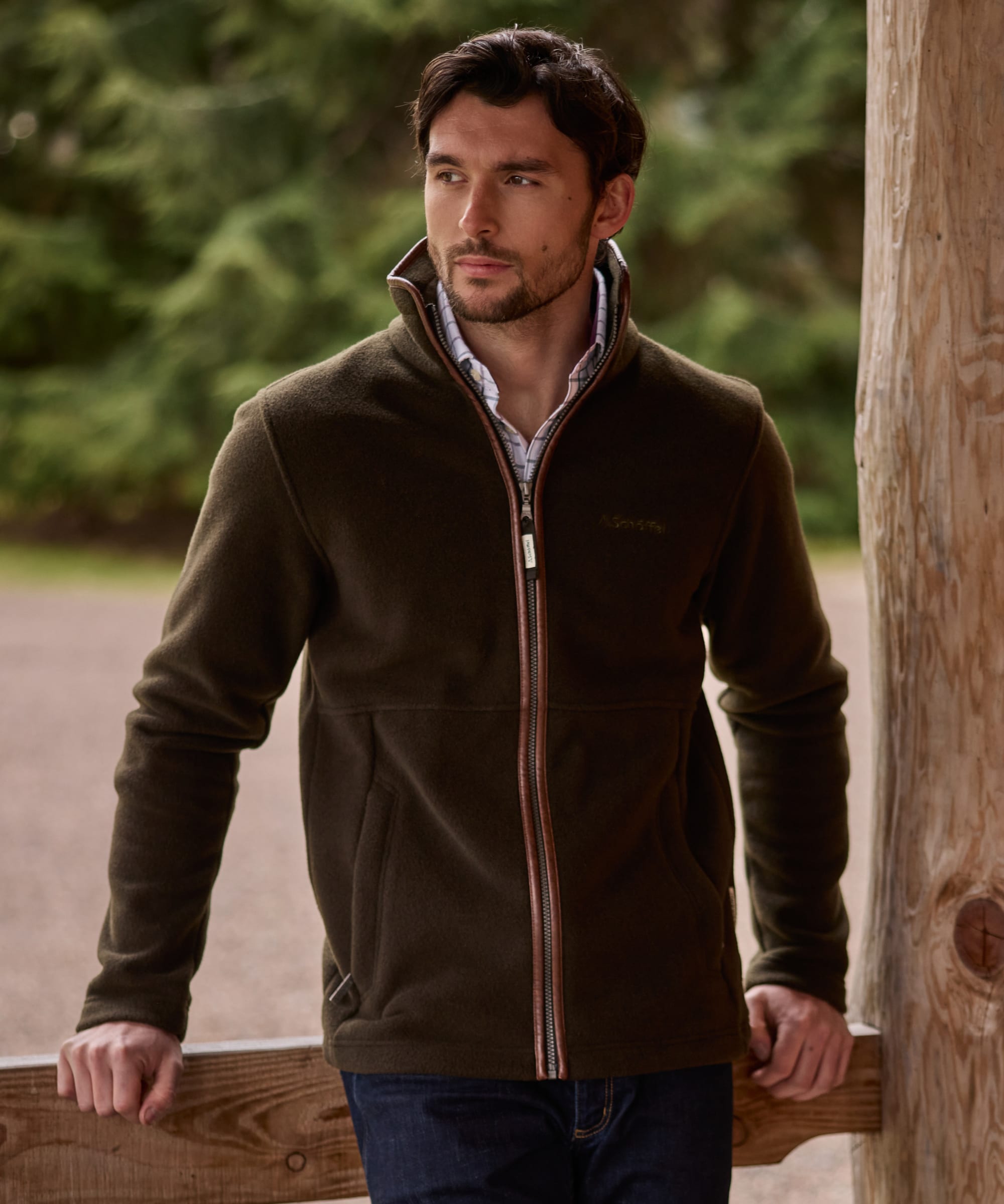 A man wearing a dark brown fleece zip-up jacket over a collared shirt and jeans, standing outdoors next to a wooden post.