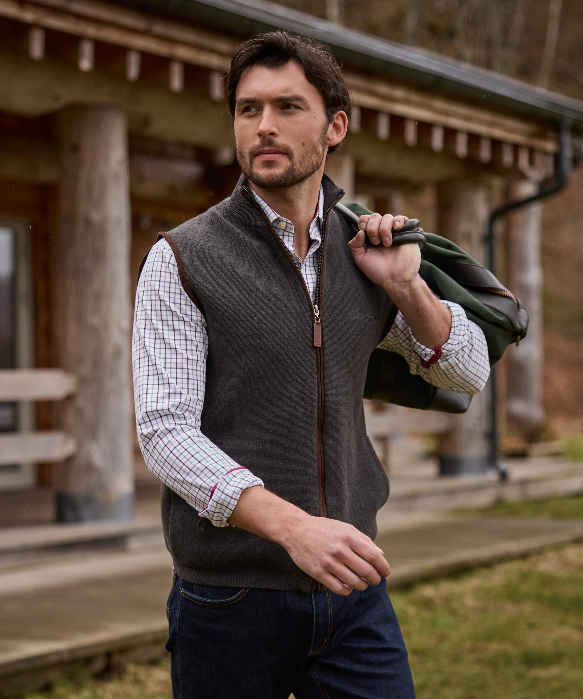 A man wearing a dark grey zip-up fleece gilet  over a white and red checked shirt, with dark blue jeans and a green backpack slung over his shoulder, standing in front of a rustic wooden cabin.