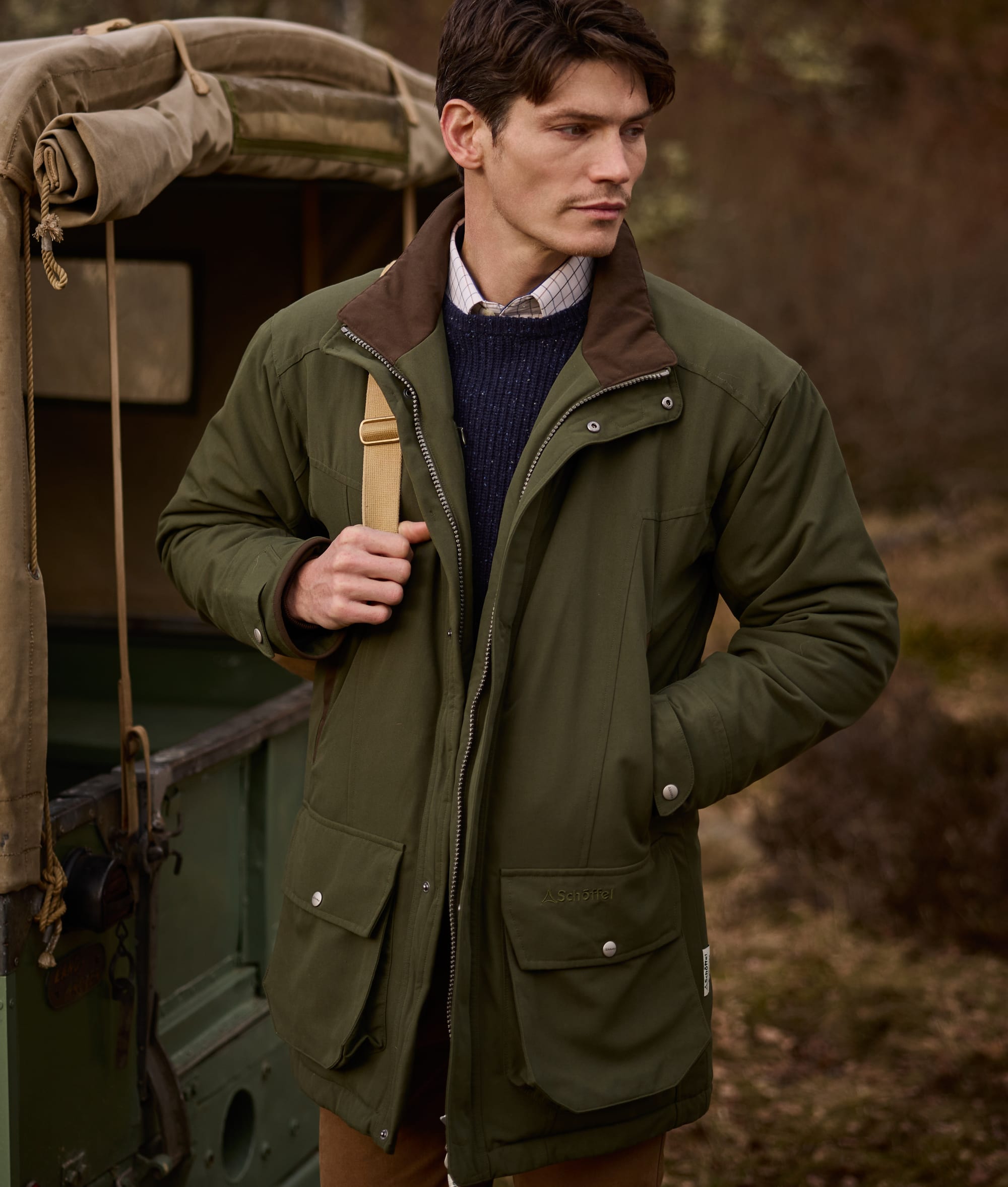 A man wearing an olive green outdoor jacket with brown collar trim, standing outdoors next to a vintage green vehicle.