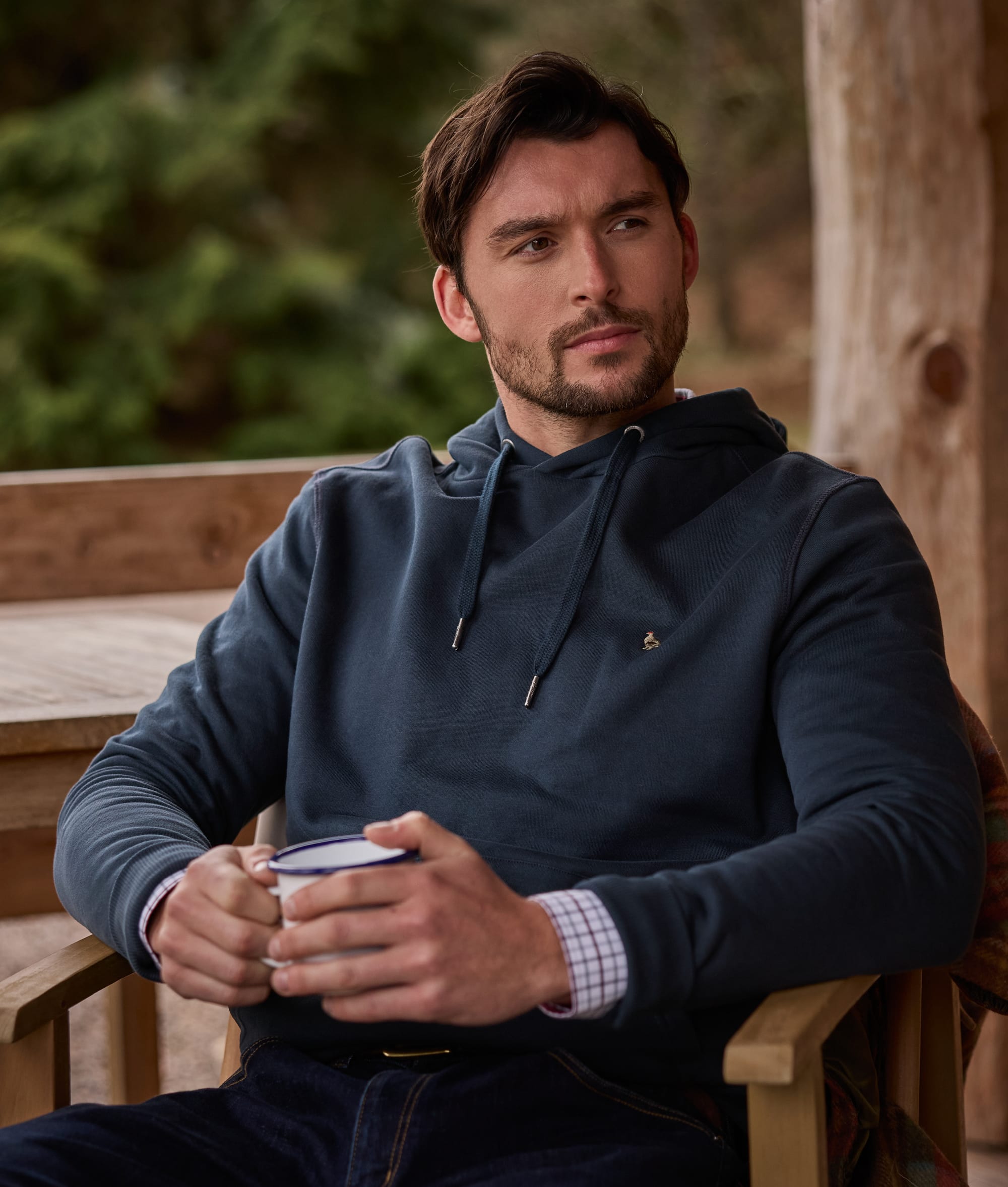 A man wearing a dark slate blue hoodie sits in a wooden chair on a porch, holding a mug.