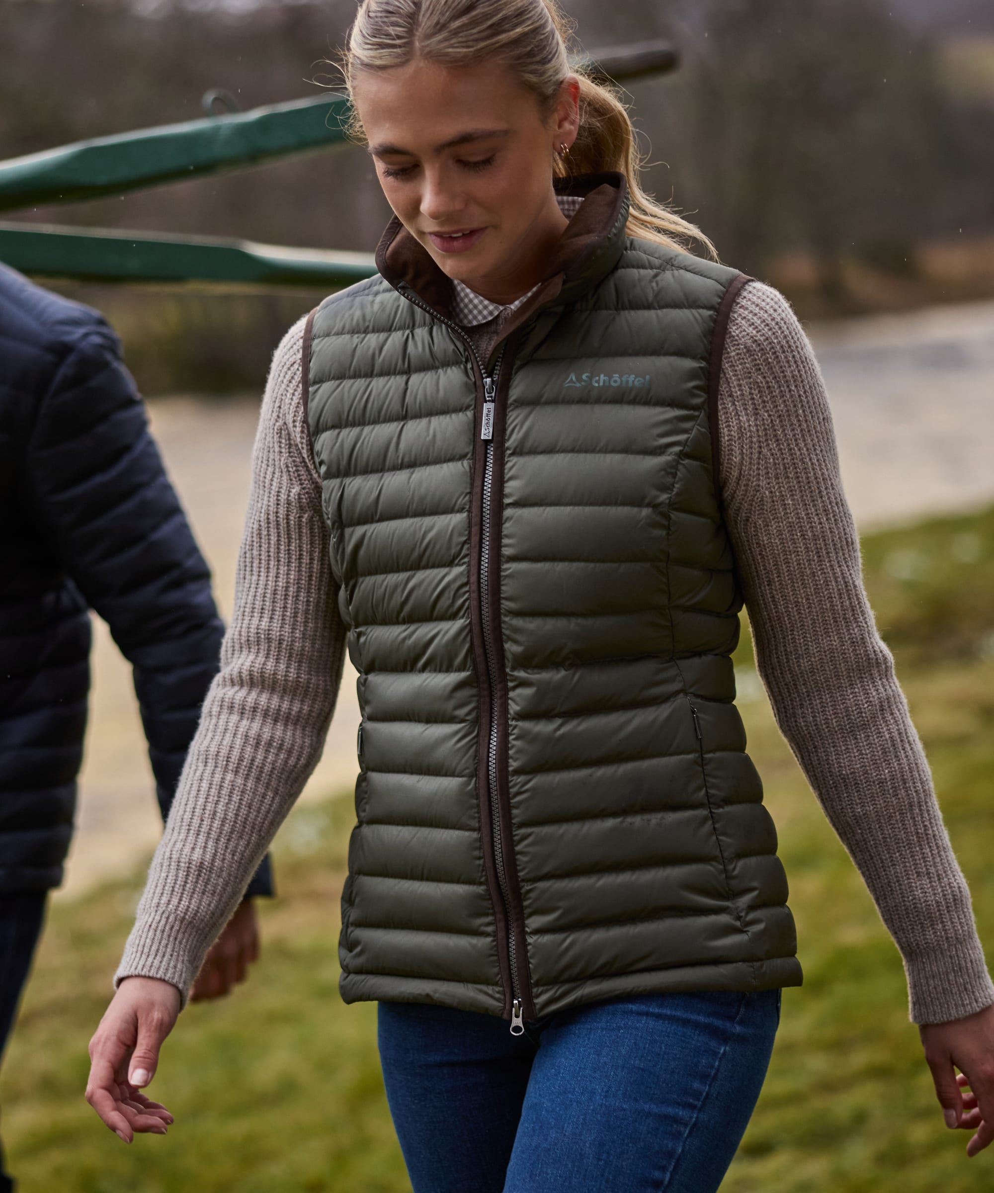 A woman wearing an olive green quilted puffer gilet over a beige ribbed fisherman jumper and blue jeans, walking outdoors in a grassy area.