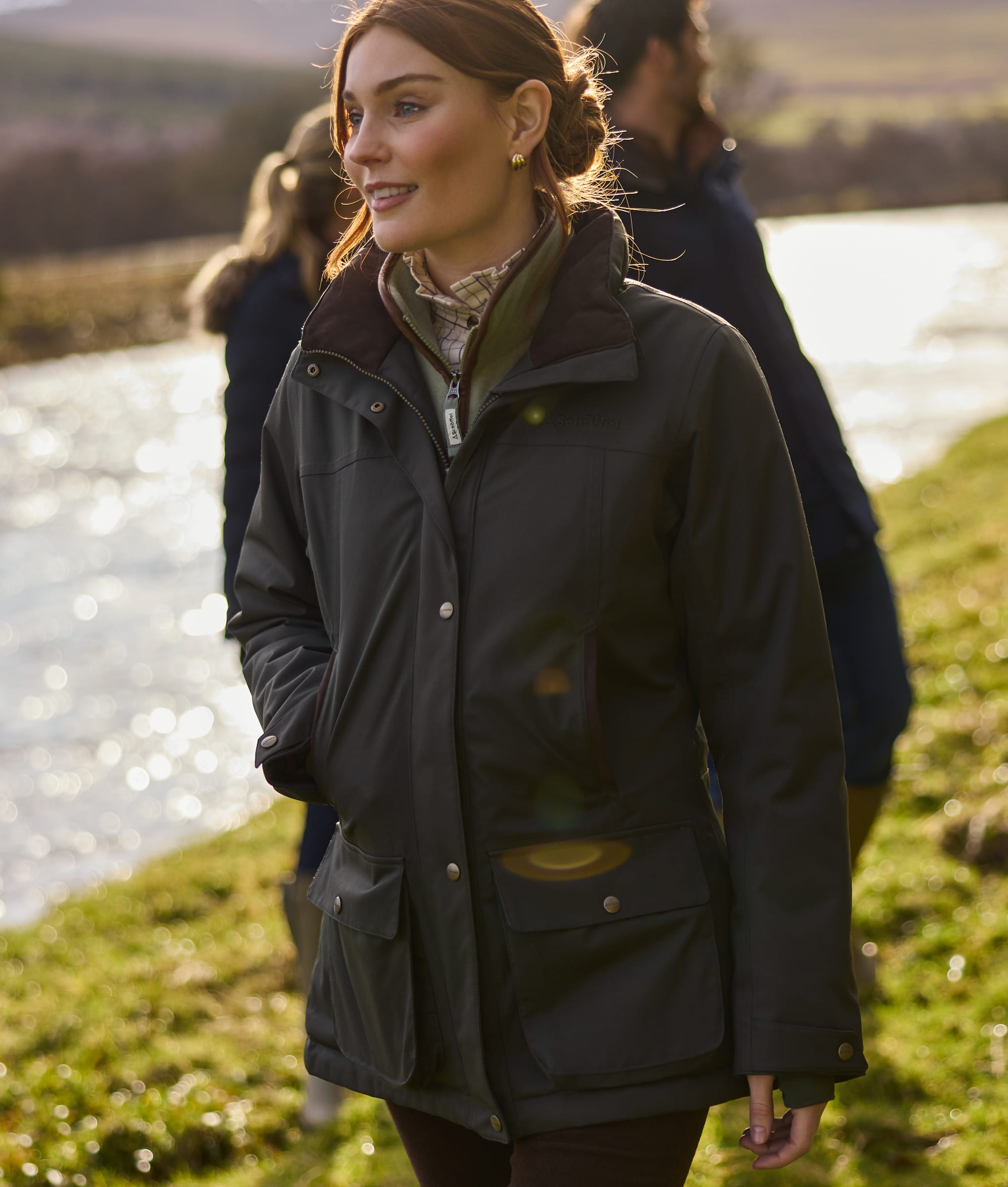 Woman wearing dark green outdoor jacket with a brown collar stands outdoors near a river