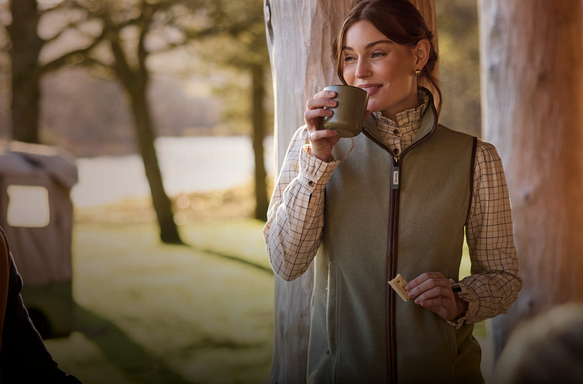 A woman outdoors by a tree drinking from a mug, wearing a green gilet over a plaid shirt.