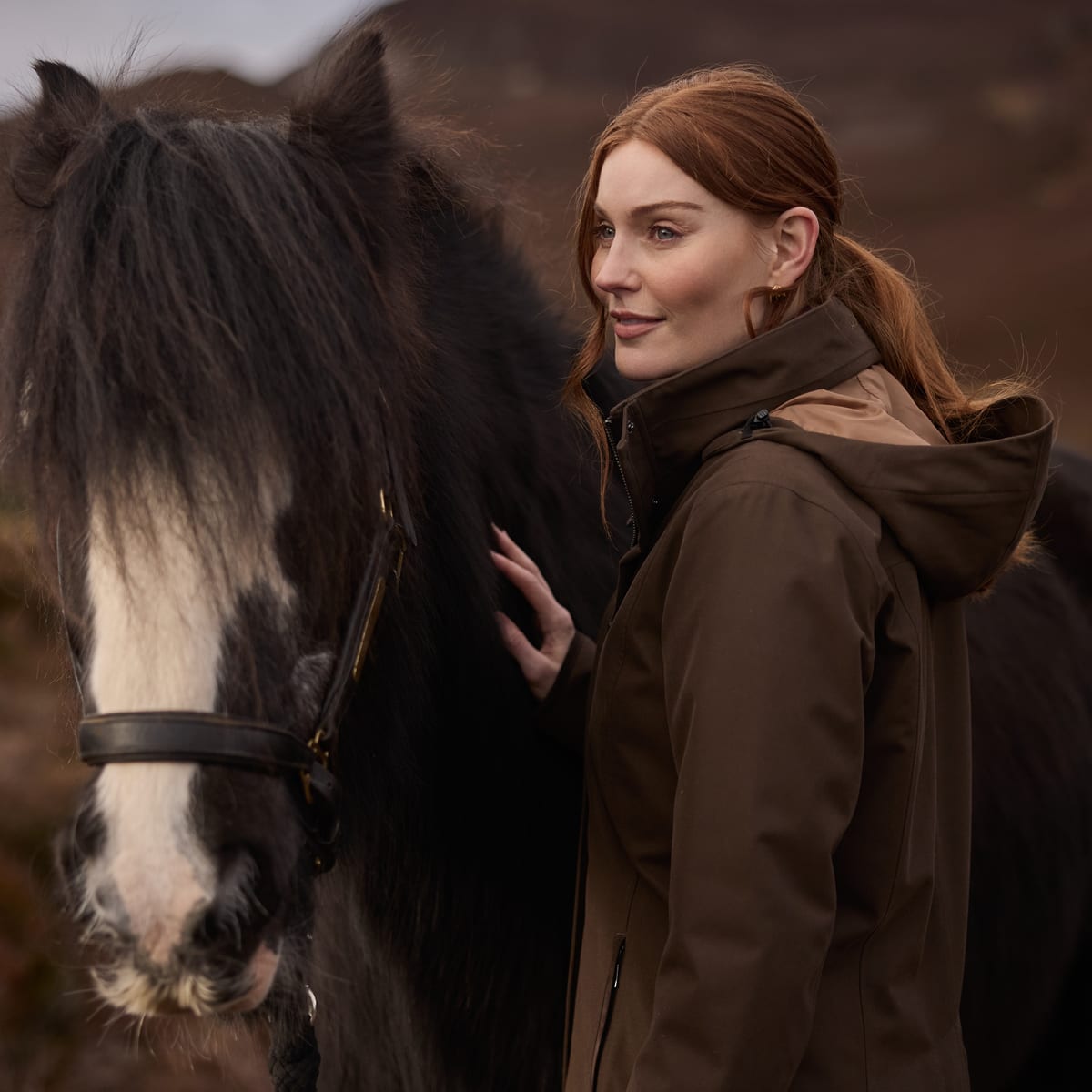 A woman with red hair stands beside a large black horse outdoors, wearing a brown hooded