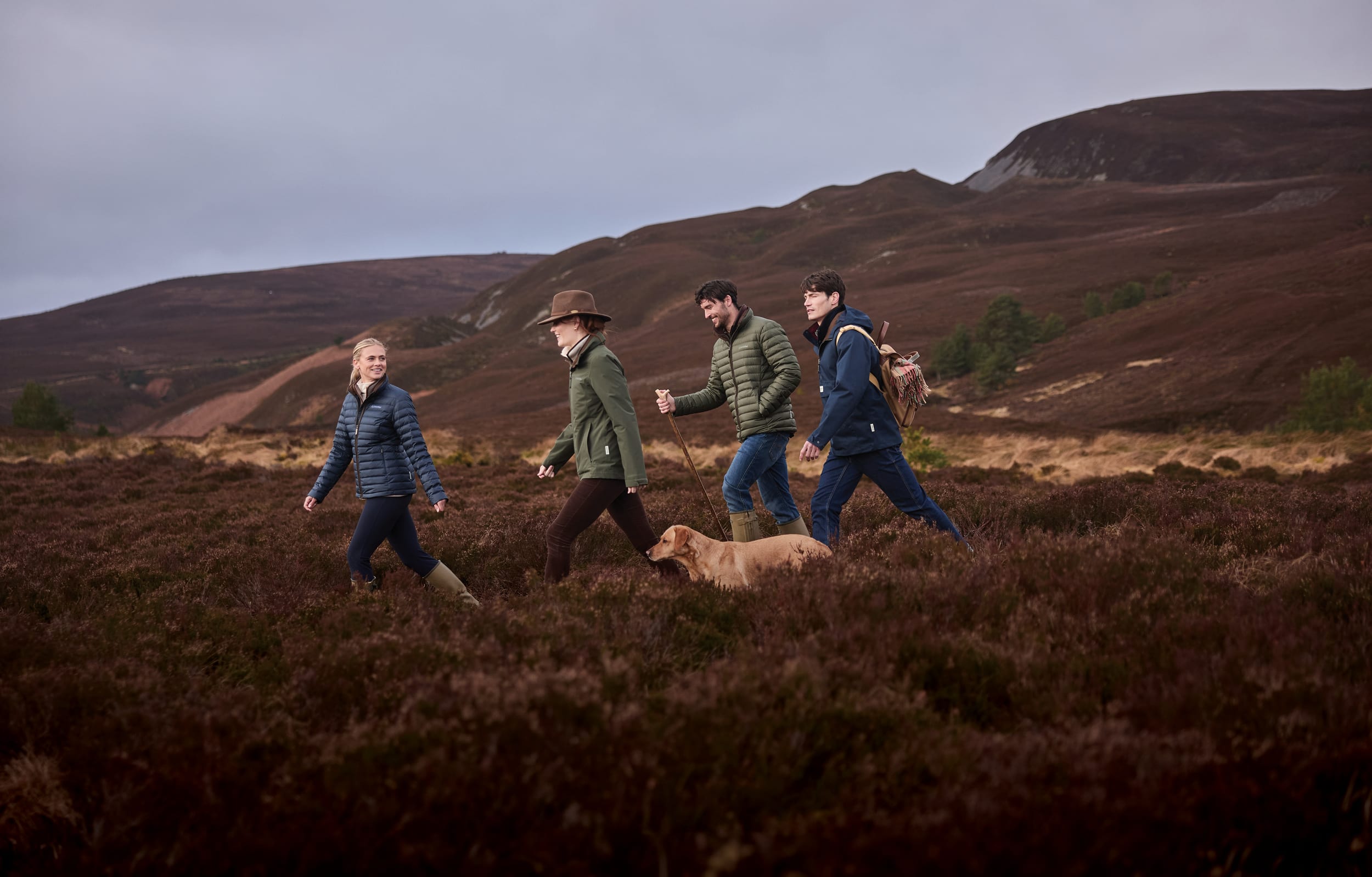 Four people walking across a moorland landscape with rolling hills in the background, accompanied by a dog. They are dressed in outdoor jackets and boots.