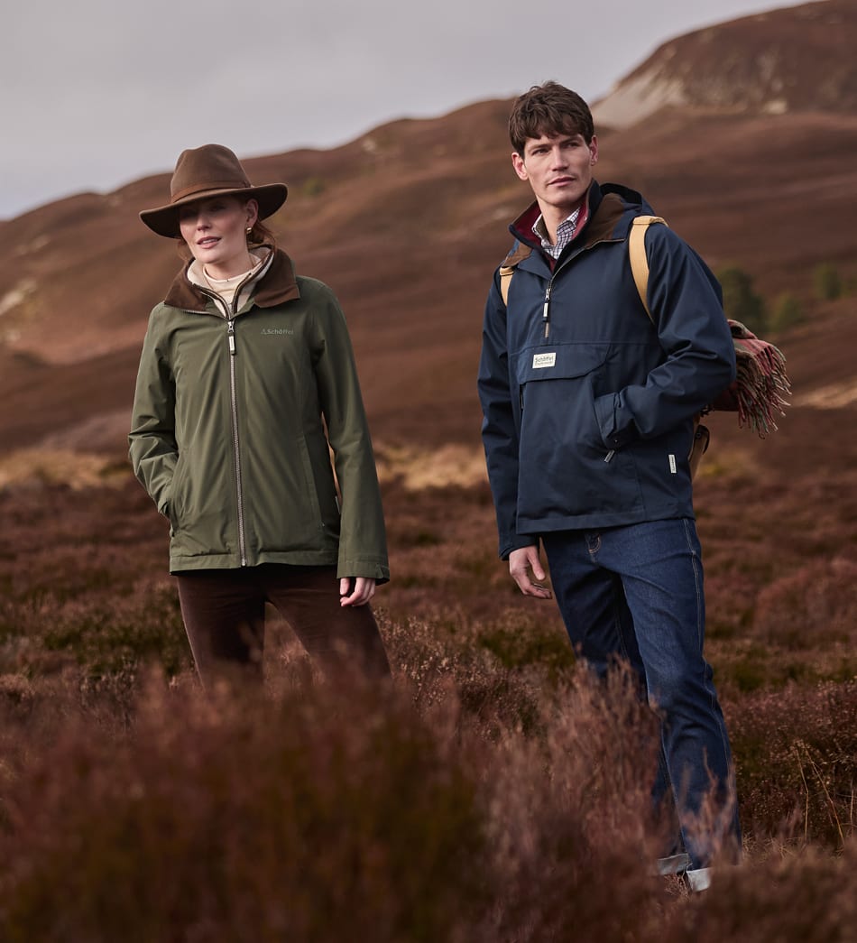A man and a woman standing in a moorland landscape wearing outdoor jackets. The woman on the left wears an olive green zip-up jacket and a brown wide-brim hat, while the man on the right wears a navy blue anorak-style jacket.