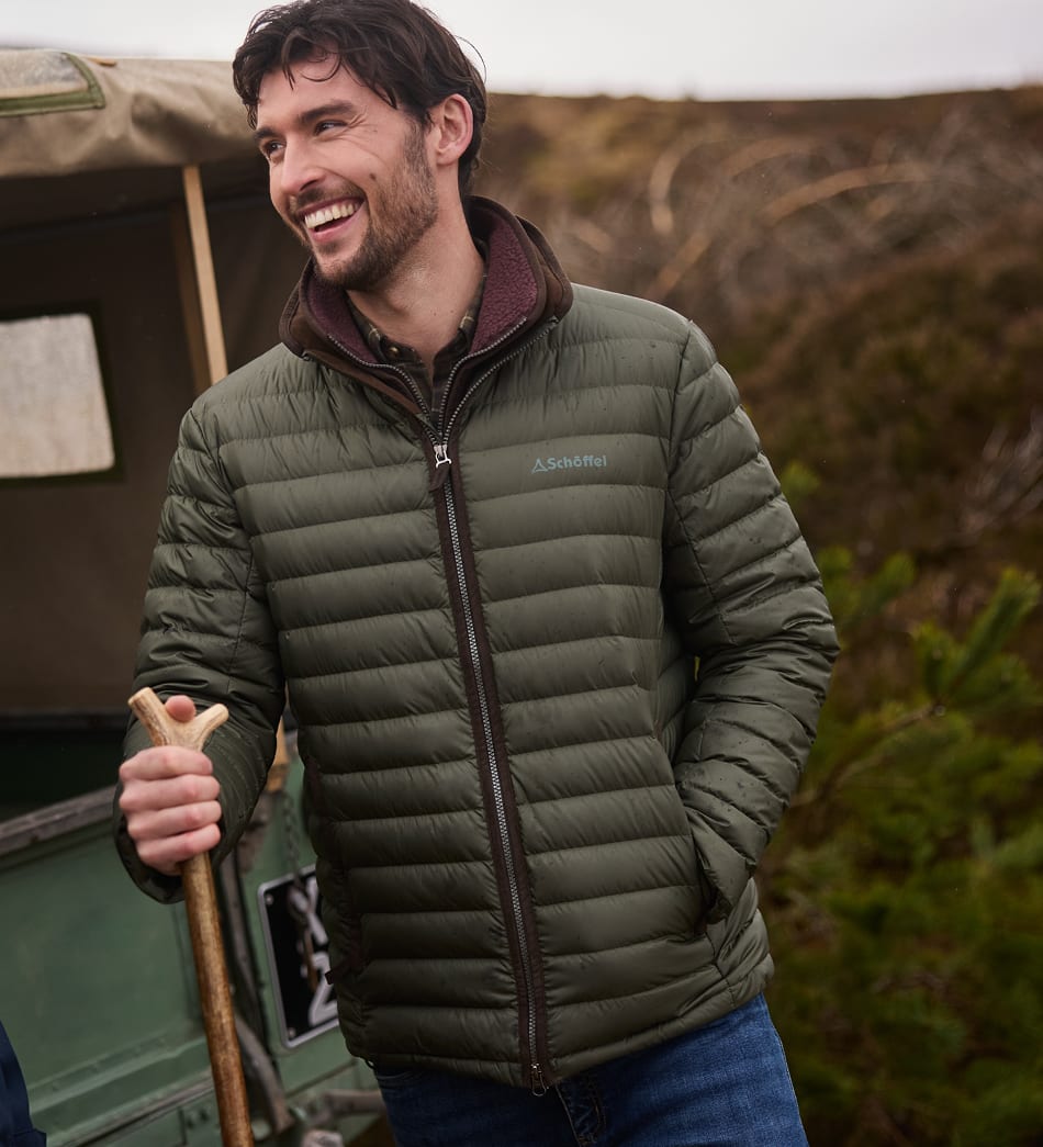 A man wearing an olive green puffer jacket with a dark brown zipper standing outdoors next to a vintage green vehicle.