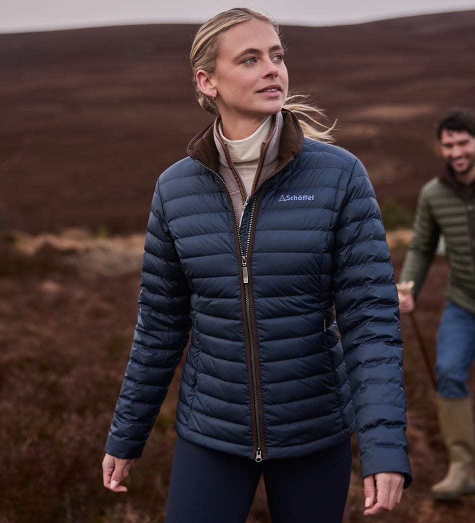 A woman wearing a navy blue insulated puffer jacket with brown collar trim stands in an outdoor moorland setting, with a man blurred in the background.