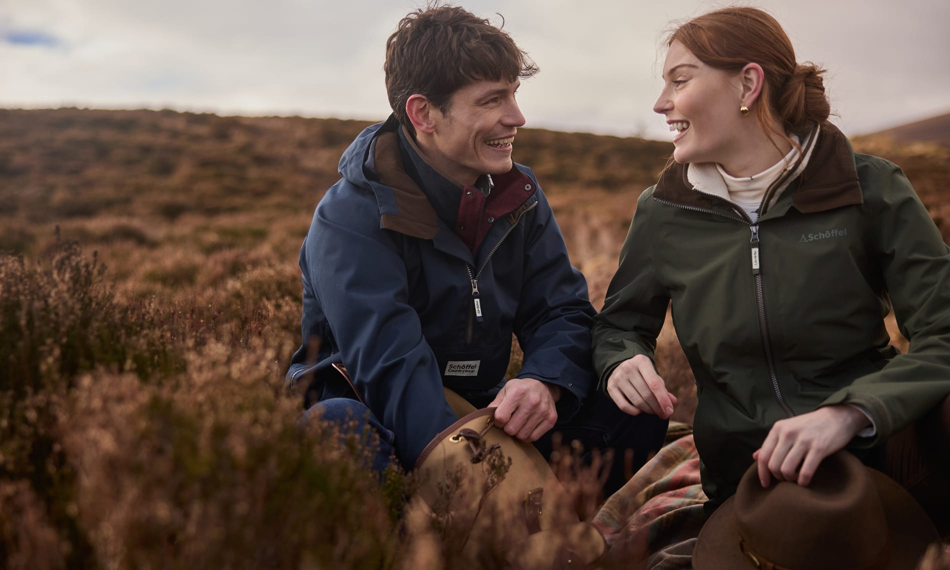A man and a woman sitting outdoors in a grassy, hilly landscape, smiling at each other. The man wears a blue hooded jacket, and the woman wears a green waterproof jacket.