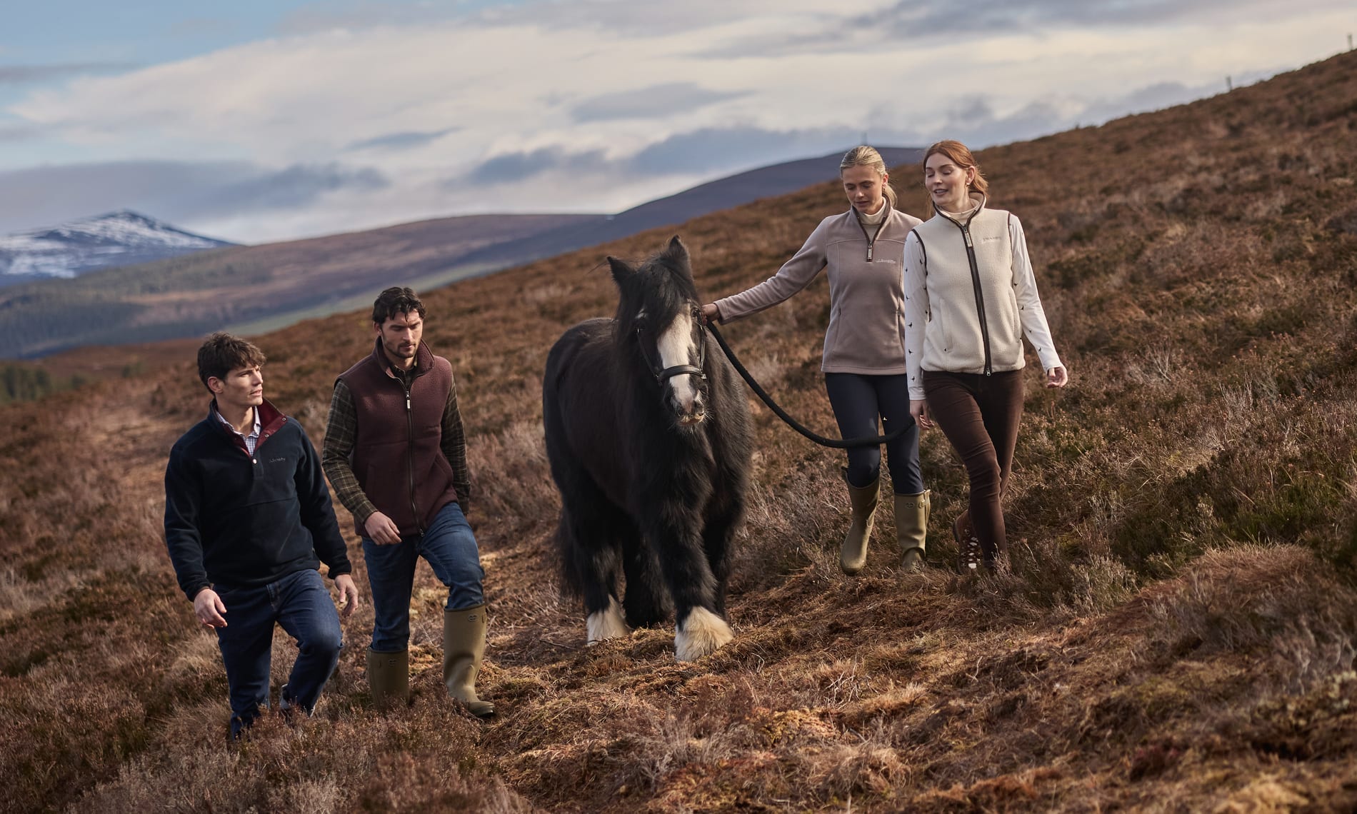 Four people walking with a large black horse across a rugged brown hillside in an outdoor mountain landscape under a cloudy sky.