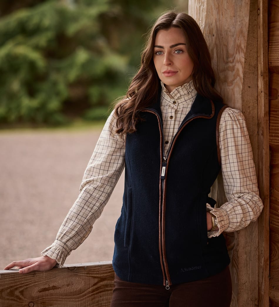 A woman wearing a dark navy gilet over a beige checked shirt leans against a rustic wooden post outdoors.