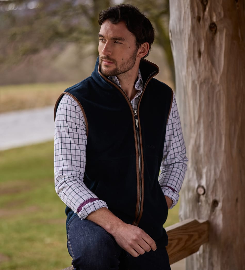 A man wearing a dark navy fleece gilet over a white and blue checked shirt, sitting outdoors.