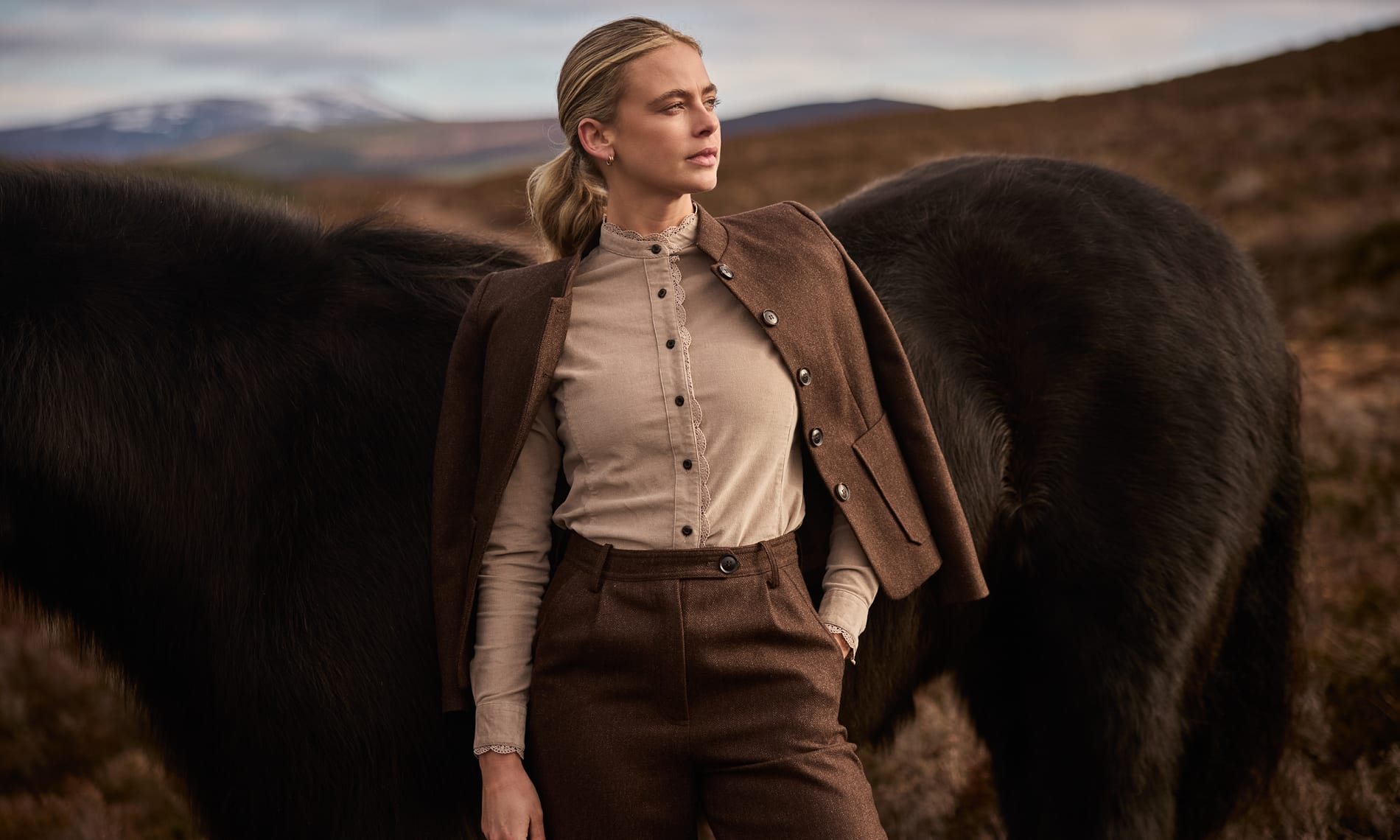 A woman stands outdoors in a rugged landscape against a dark horse wearing a beige high-neck blouse, brown tweed tailored jacket and trousers.