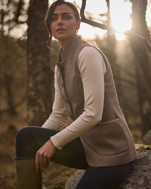 A woman wearing a tweed vest over a cream ribbed long-sleeve top and dark leggings, sitting outdoors on a rock in a sunlit forest.